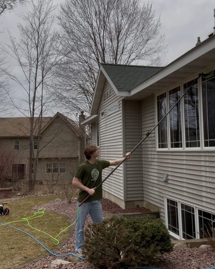 Young man using a long pole to clean windows on a house surrounded by trees and grass.